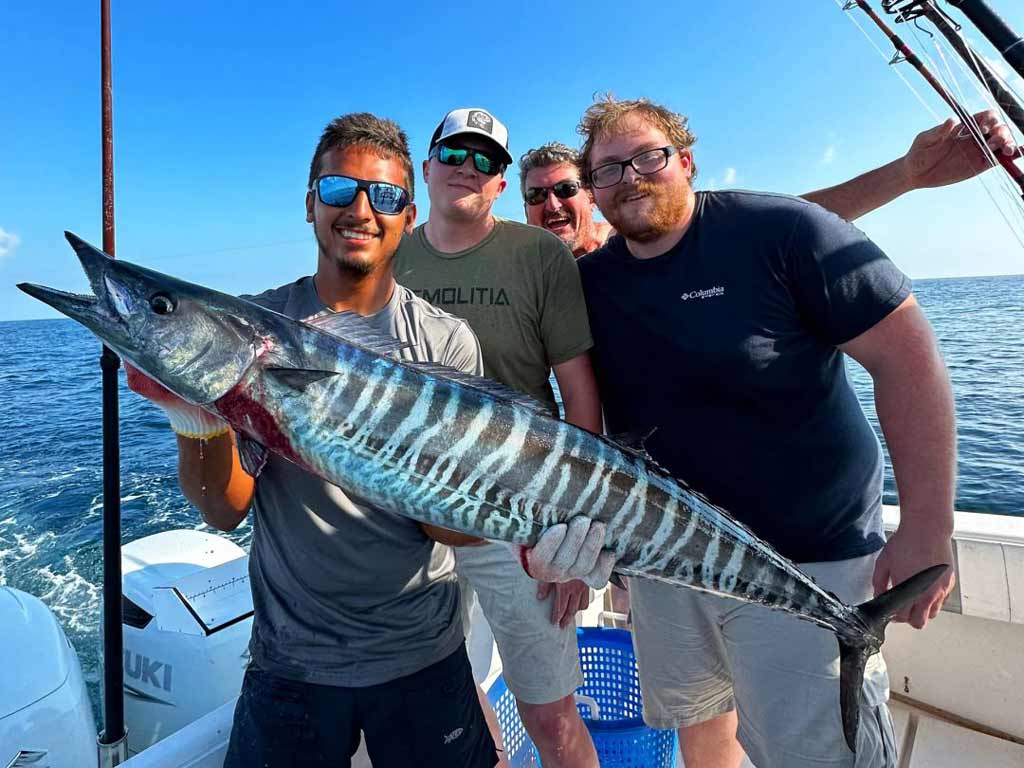 Four anglers are standing on a charter boat, gathered around a massive Wahoo to pose for the camera on their Alabama deep sea fishing trip
