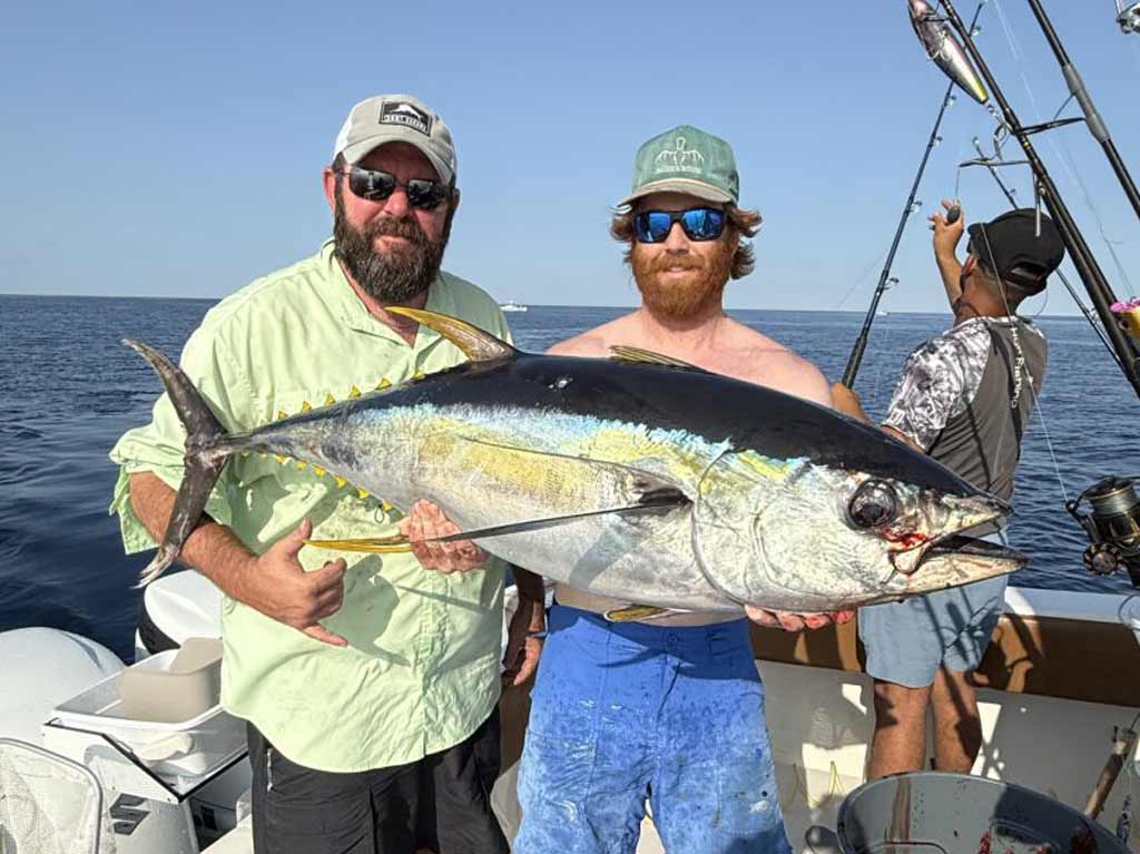 Two anglers on a boat hold a large Yellowfin Tuna, while another person fishes in the background during their offshore fishing trip out of Orange Beach, Alabama. The ocean and blue sky are visible.