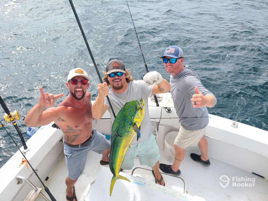 Three anglers on a boat pose with a large Mahi-Mahi, grinning after their deep sea fishing trip, with the ocean sparkling behind them.