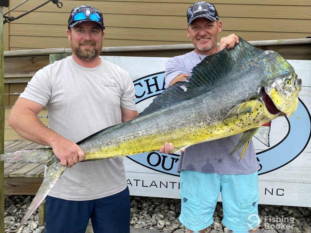 Two proud anglers stand outside holding a large Dolphinfish, also known as Mahi-Mahi, posing in front of a sign and wooden wall North Carolina’s dock
