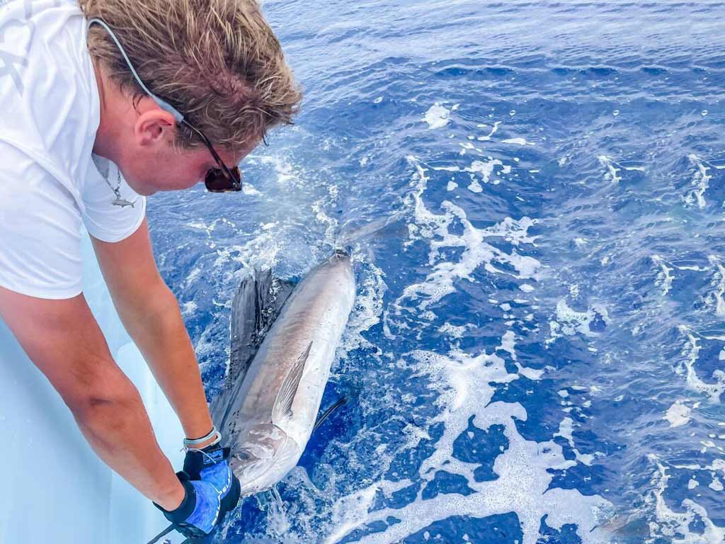 An angler in sunglasses and gloves holds a big Marlin, a catch worthy of the Big Rock Blue Marlin Tournament, beside a boat on bright blue ocean water.