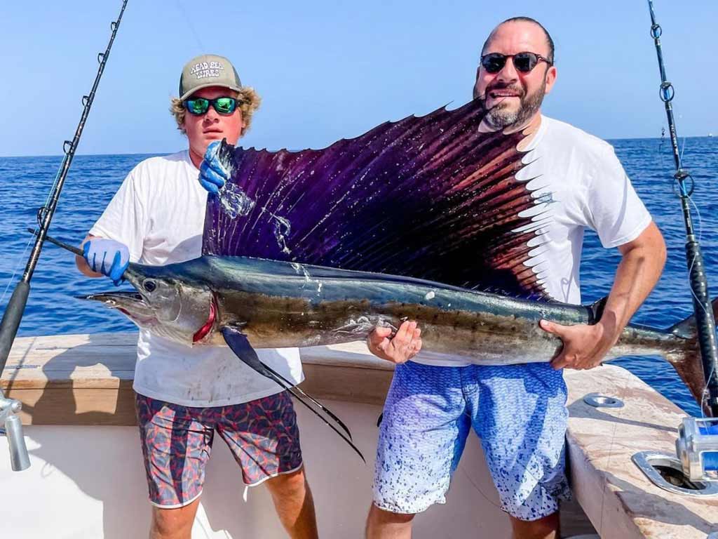 Two proud anglers on a boat are holding a large Sailfish with a dark purple, sail-like dorsal fin; the ocean’s in the background at the deep sea fishing trip in North Carolina