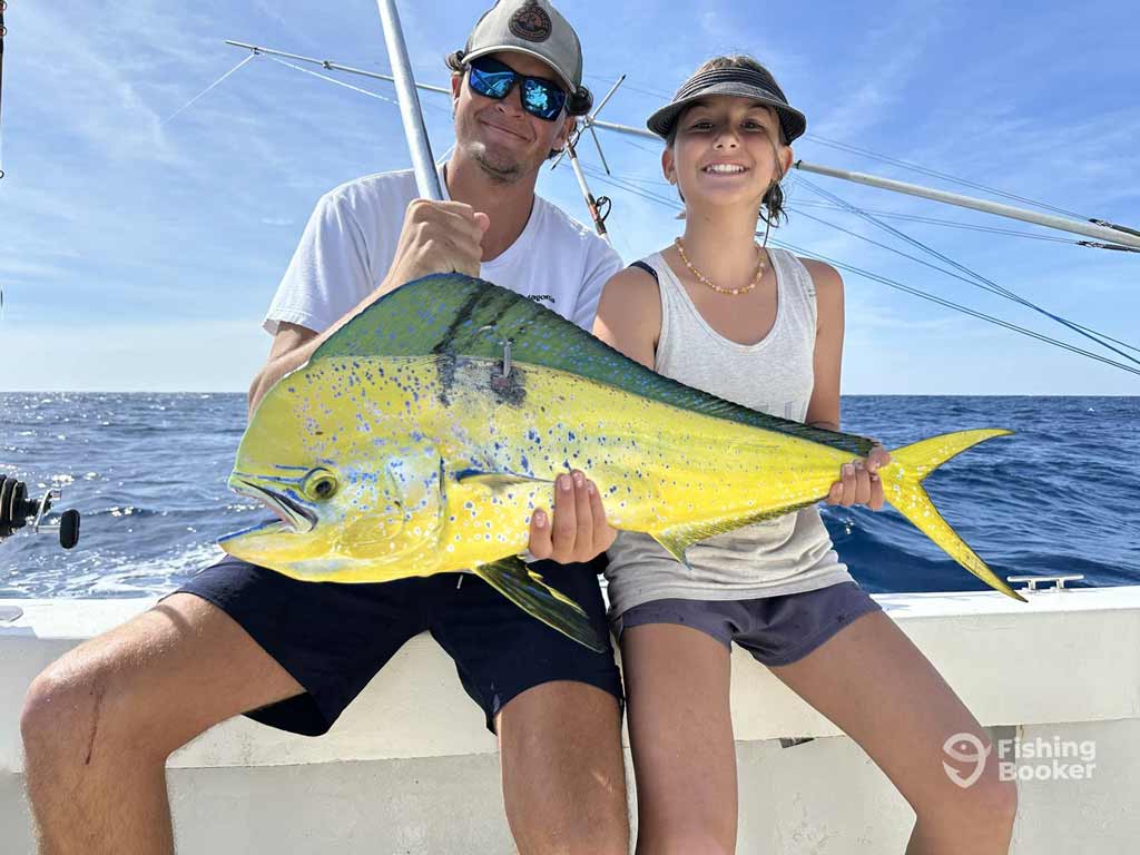 Two anglers sit on a boat holding a colorful Mahi-Mahi, a catch worthy of the Big Rock Blue Marlin Tournament, with fishing rods and the ocean below a clear blue sky.