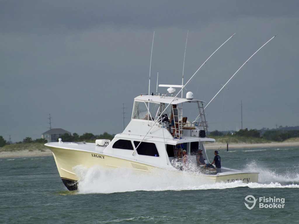 A white fishing boat named “Legacy” speeds near the shore under cloudy skies, with two people on board, possibly heading to the North Carolina’s Crystal Coast