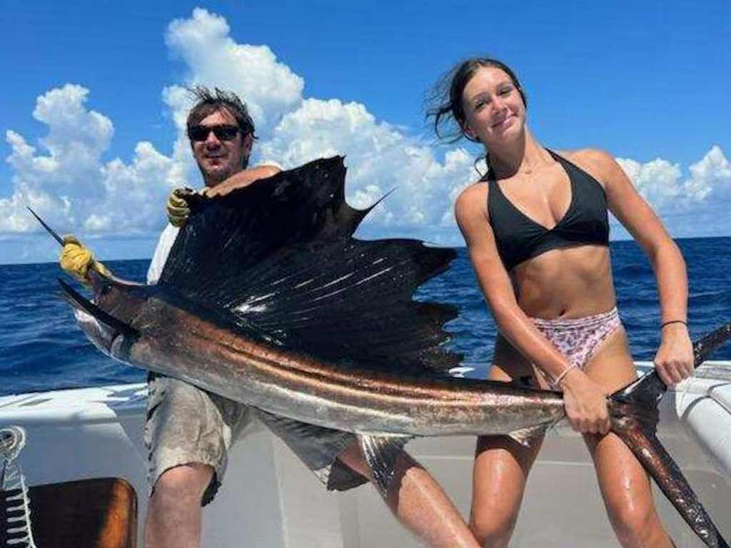 Man and woman on a boat holding a large Sailfish under a blue sky with clouds—it’s reminiscent of trophy catches at the Big Rock Blue Marlin Tournament.