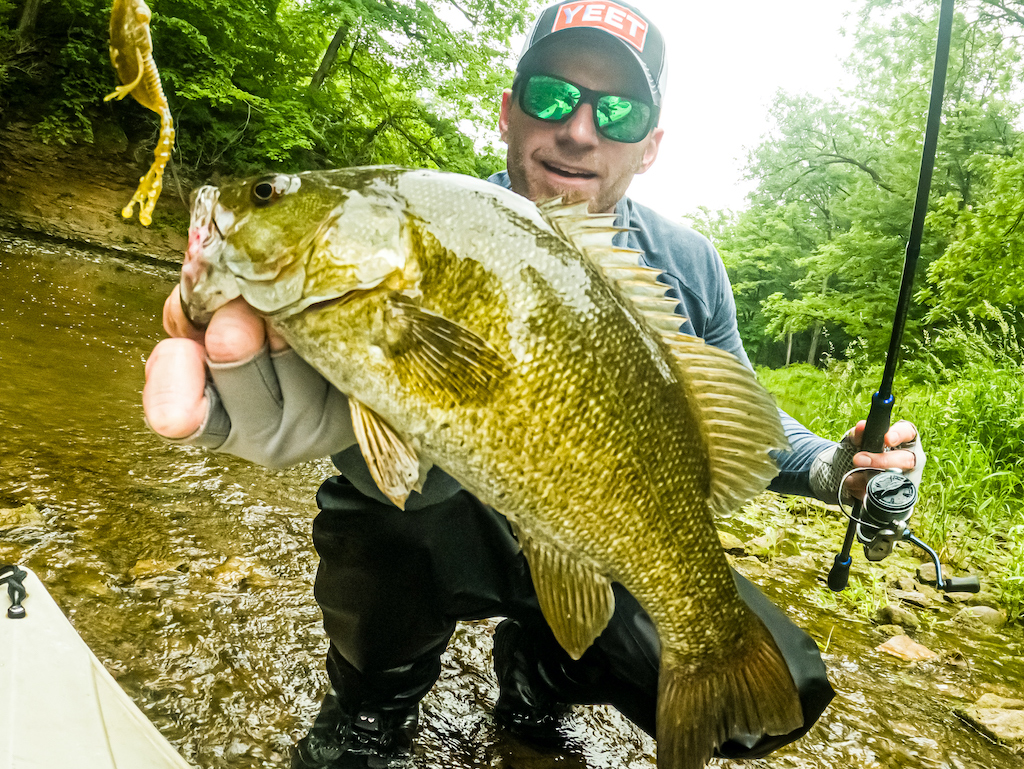 A man holding a big Smallmouth Bass close to the camera by a stream, wearing sunglasses and a cap, with a fishing rod in the other hand.
