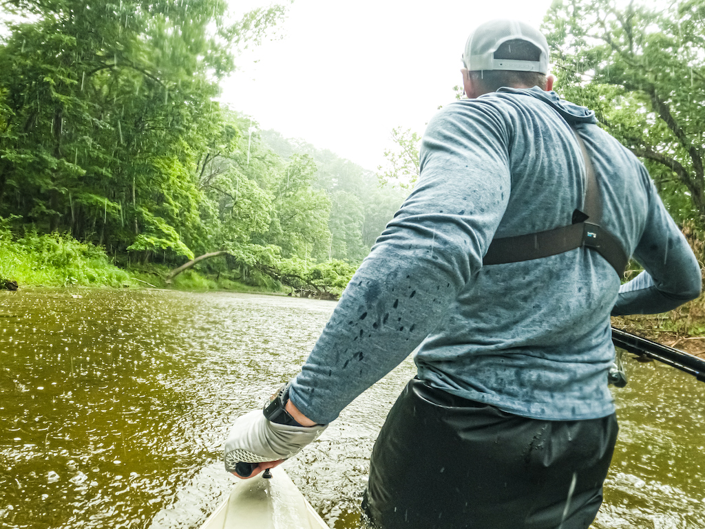 A view from behind of a man wearing a blue long-sleeve shirt and cap as they paddle a canoe down a river, hoping to catch a big Smallmouth Bass in the dense green forest on a cloudy day.