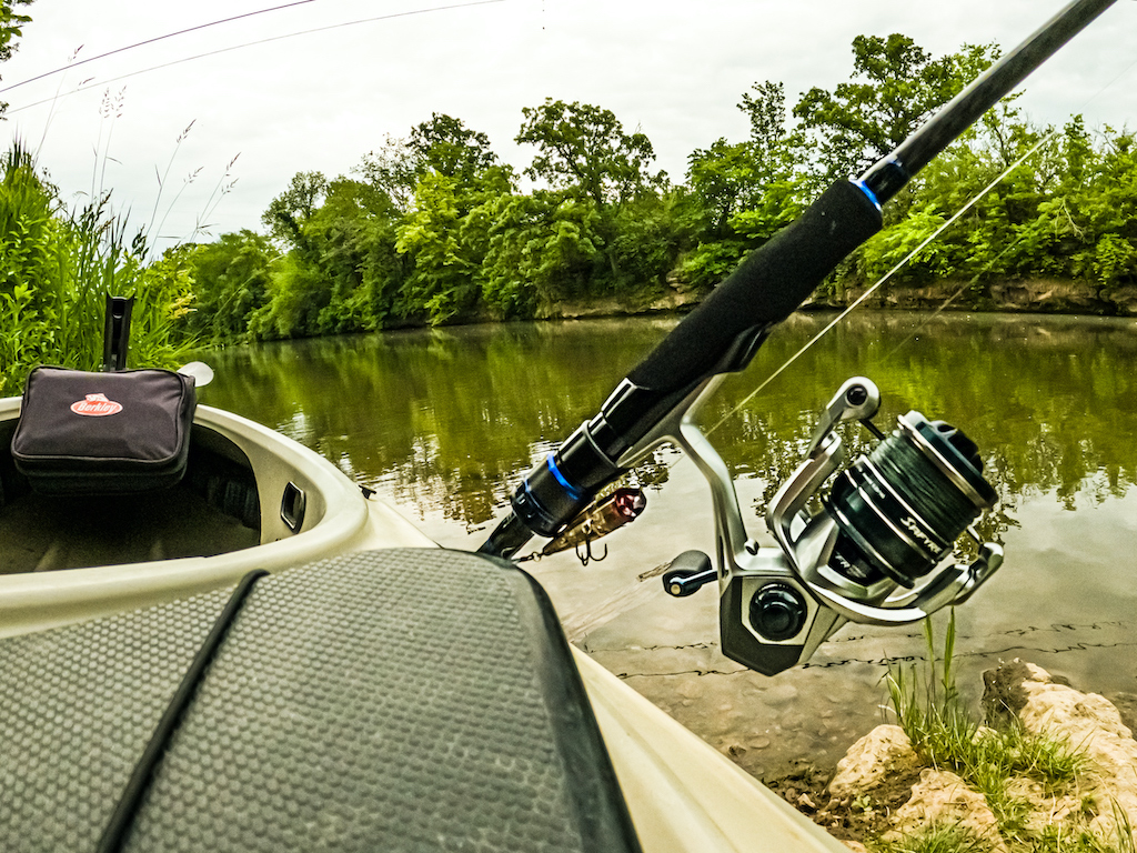 A fishing rod resting on the side of a boat by a calm riverbank, surrounded by green trees.