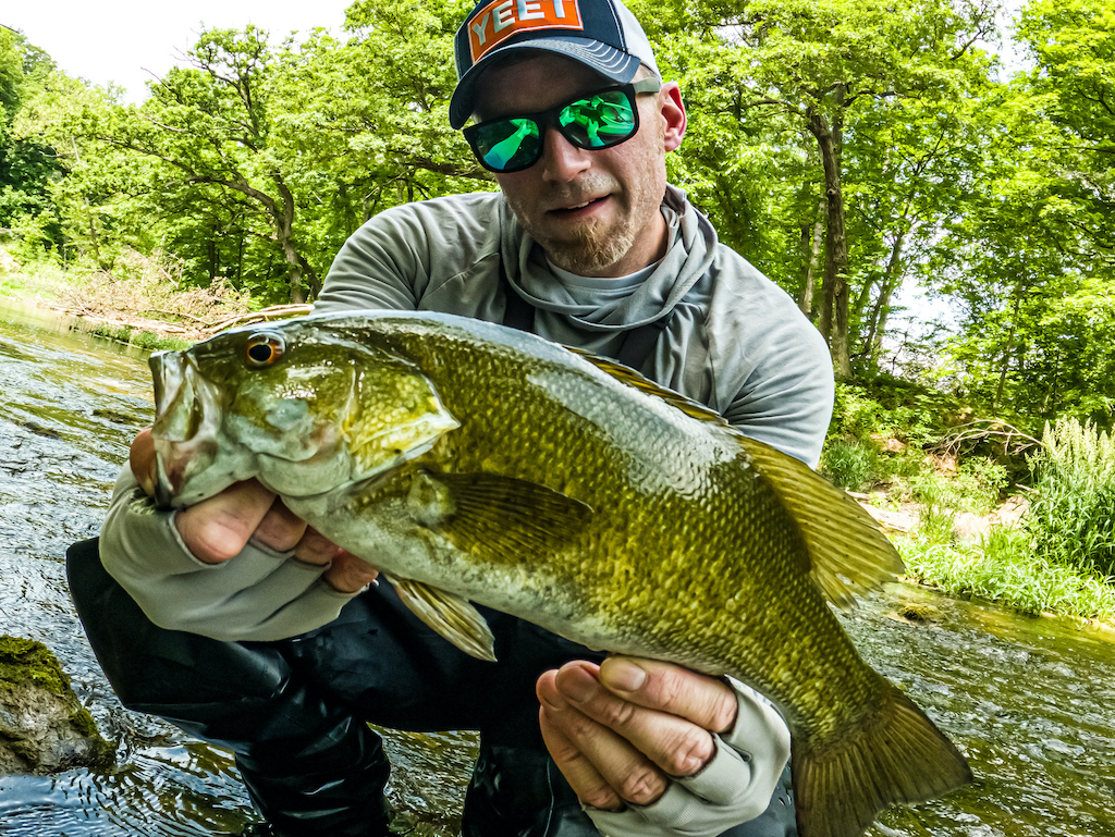 A man wearing sunglasses and a cap, holding a big Smallmouth Bass near a stream with green trees and water in the background.