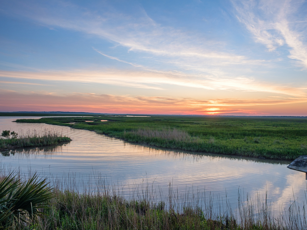 You’re looking at a calm marsh at sunset, with still water, lush green grass, and a sky full of soft clouds painted in bright colors.