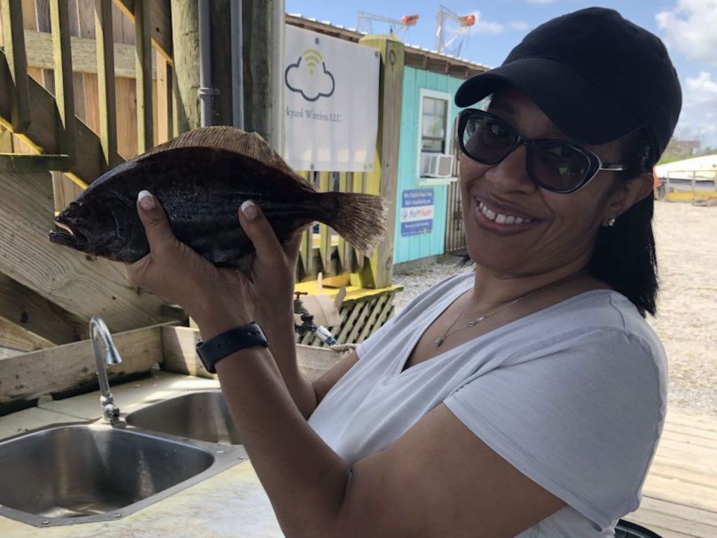 A woman wearing sunglasses and a cap as she holds up a Flounder near an outdoor sink, smiling at the camera with buildings and a cloudy sky in the background.