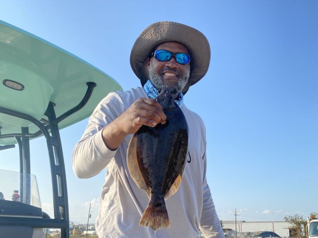 A man in a hat and sunglasses smiles while holding up a Flounder next to a boat under a clear sky.